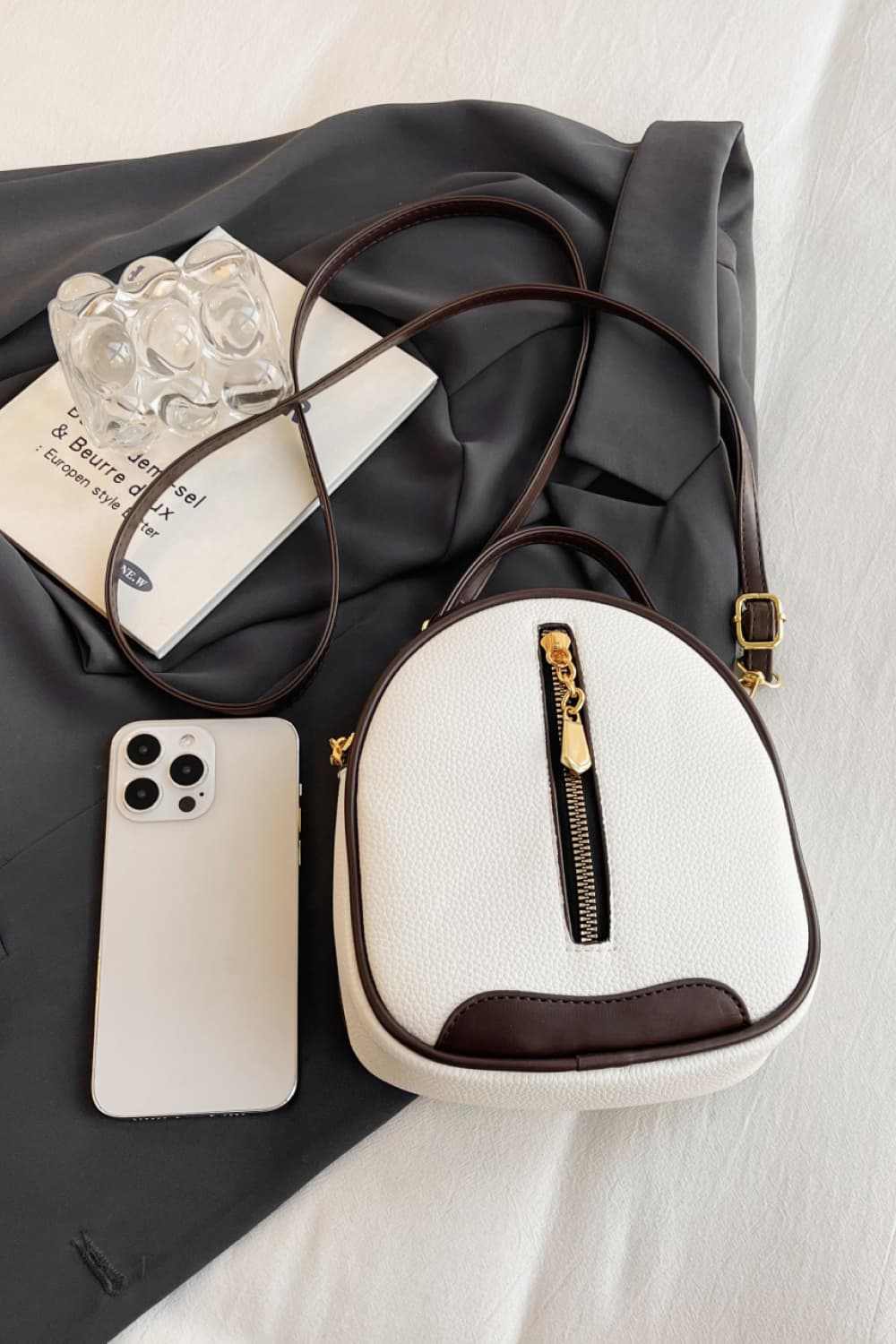 Small PU leather handbag with gold zipper and adjustable strap, displayed with a smartphone and fabric background.