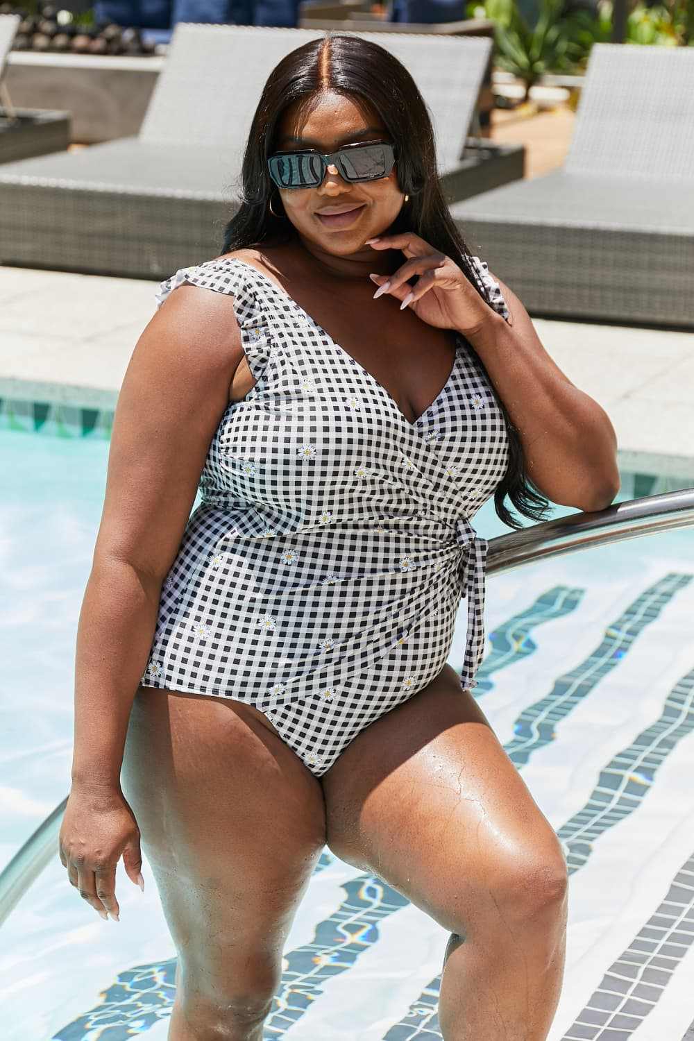 Plus-size model in black Marina West Swim one-piece swimsuit with ruffle faux wrap design by pool.