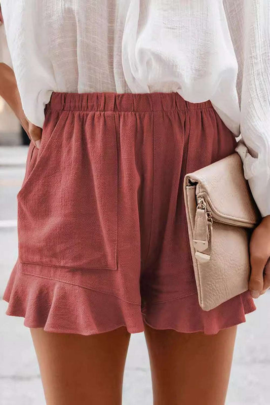 Full size ruffled elastic waist shorts in red, featuring pockets, worn by a model with a white blouse and holding a beige clutch.