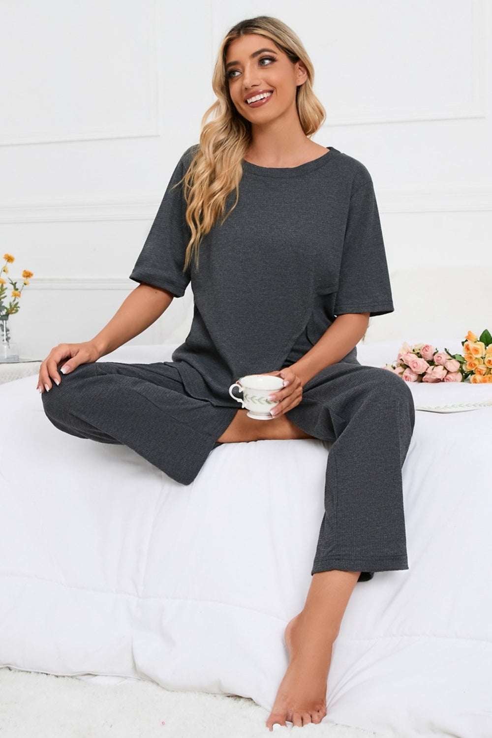 Slit Round Neck Top and Pants Lounge Set in dark color, worn by a model sitting on a bed, holding a cup.