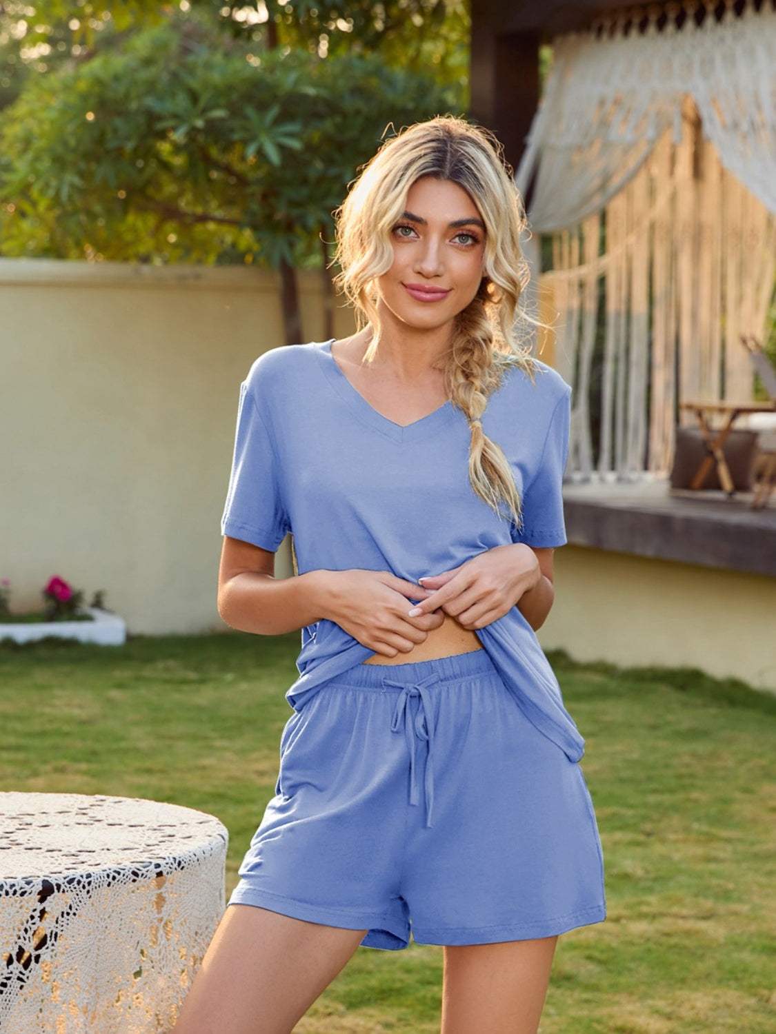 Woman wearing a light blue V-neck short sleeve top and pocketed shorts lounge set in a garden setting.