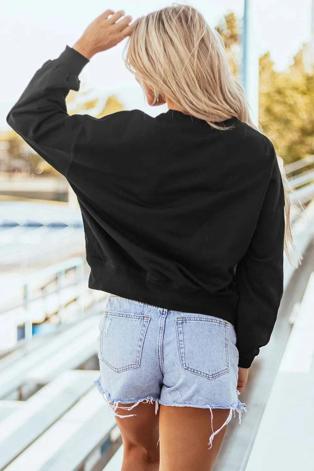 Woman wearing a black GAME DAY round neck long sleeve sweatshirt with denim shorts, standing outdoors.